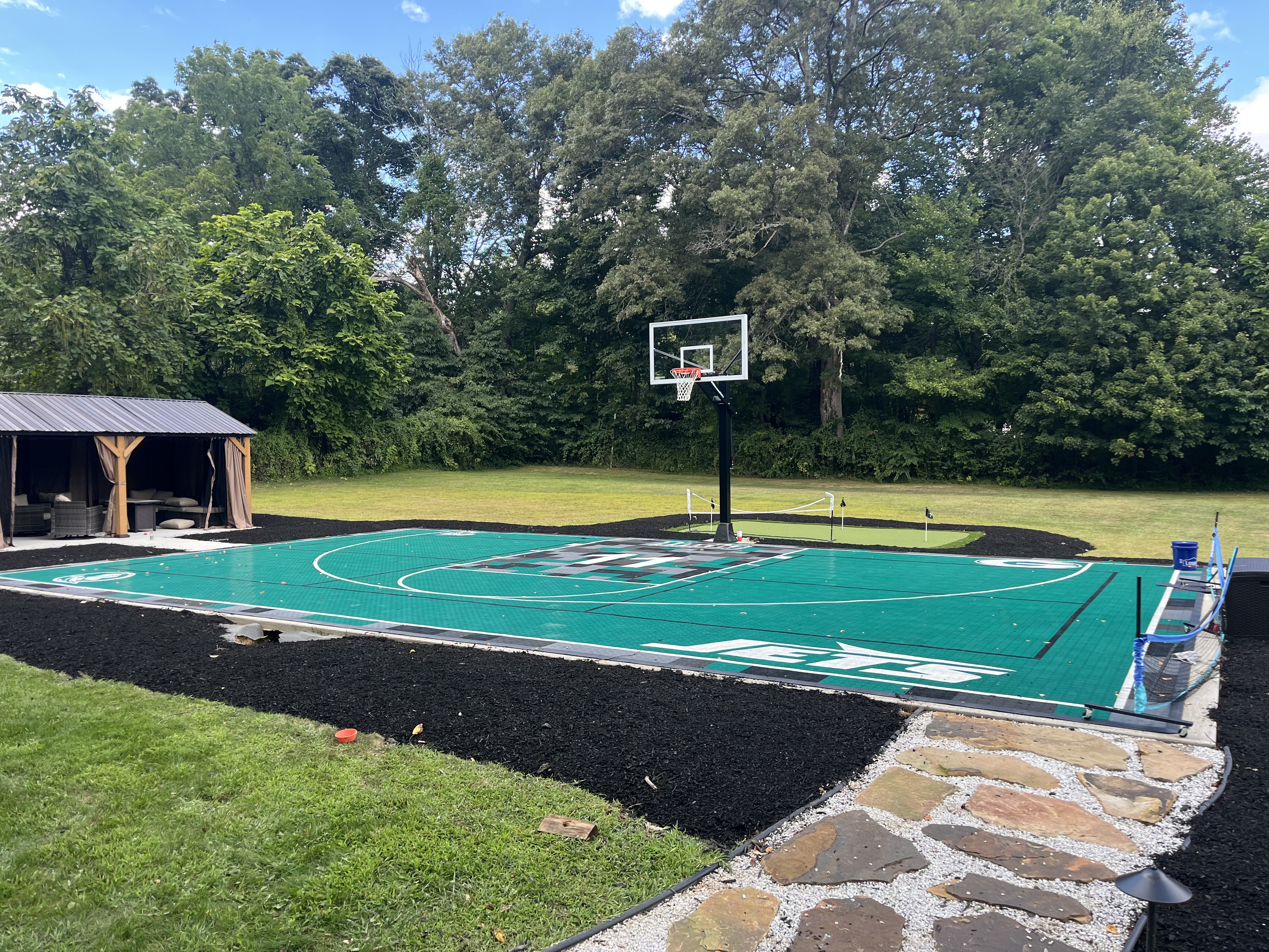 Custom backyard basketball court with green surface, rubber mulch border, gazebo, and stone walkway.