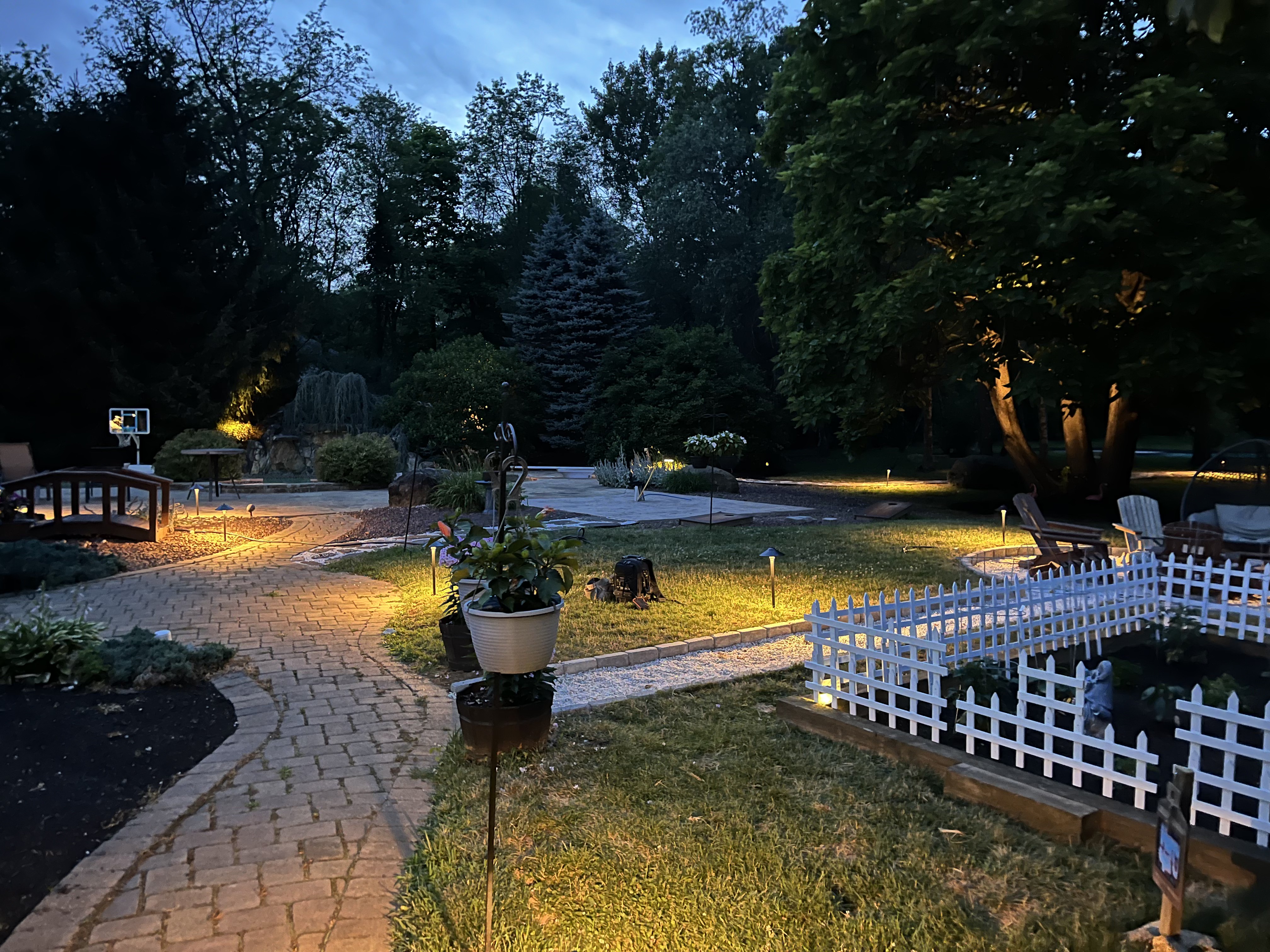 Backyard paver walkway and patio lit by landscape lighting at dusk with garden beds and fire pit.